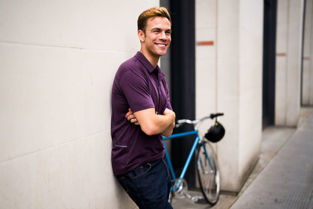 Man wearing a purple polo shirt, smiling and leaning against a wall with a blue bicycle in the background