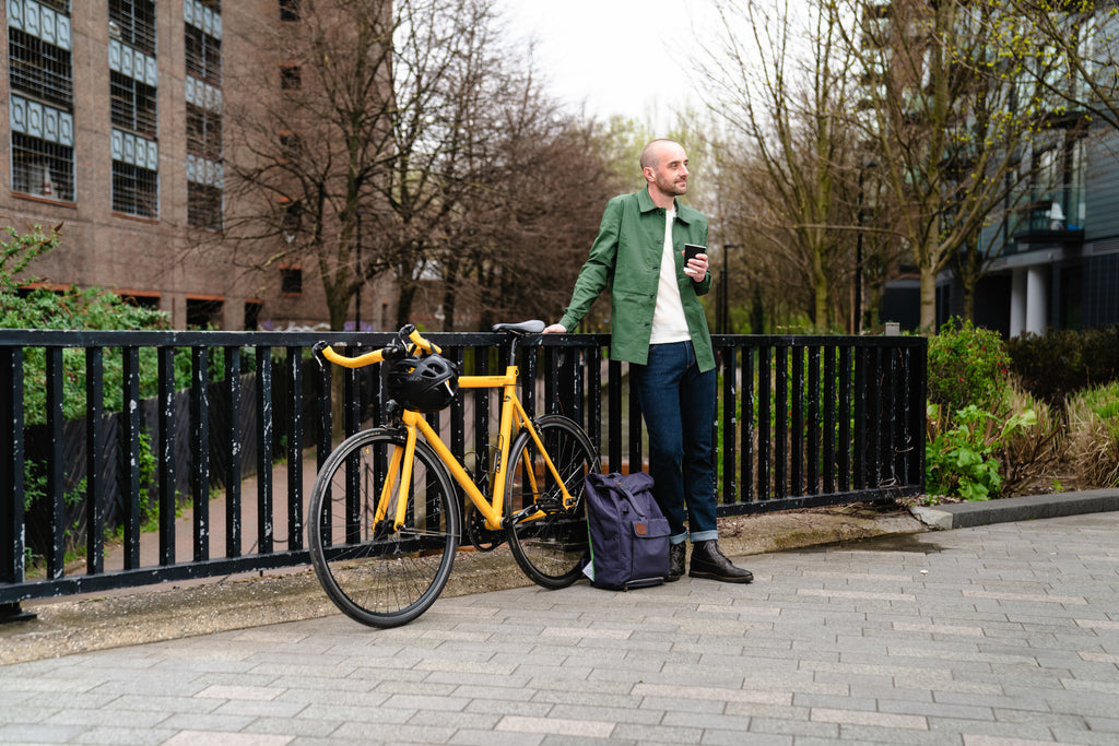 A man in green Vulpine French Workers Jacket, standing by a yellow bike with a coffee and backpack